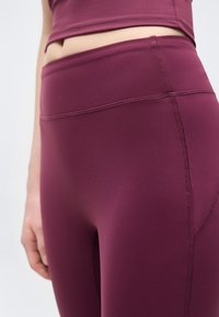 Κοντινό πλάνο ενός ατόμου που φοράει μπορντό κολάν με ψηλή μέση, χωρίς ραφές, και ταιριαστό crop top, πάνω σε απλό φόντο.