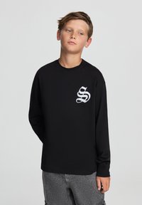 Czarny longsleeve z białym logo z przodu. Wykonany z miękkiego materiału, z okrągłym dekoltem i luźnym krojem.