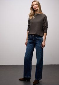 Pull marron à manches longues avec poignets côtelés, associé à un jean en denim bleu à jambes larges. Le modèle porte des bottines marron foncé. Fond neutre.