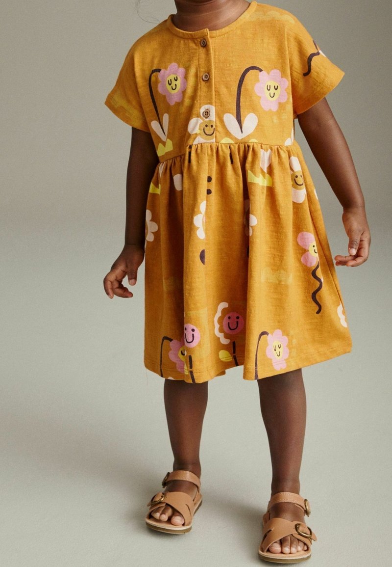 Enfant portant une robe jaune moutarde avec des motifs de fleurs souriantes et des sandales à boucle beige, debout devant un fond neutre.
