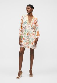 Γυναίκα που φοράει λευκό μίνι φόρεμα με μακριά μανίκια, V λαιμόκοψη, φλοράλ σχέδιο και βολάν στο τελείωμα, καθώς και μπεζ πέδιλα με ψηλό τακούνι, στέκει μπροστά σε απλό φόντο.