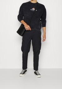 Felpa nera con girocollo e stampa "PROTEZIONE", abbinata a pantaloni cargo neri con tasche con zip e sneakers nere alte.