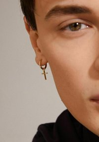 Goldene Creole mit einem kleinen Kreuzanhänger, der eine glatte metallische Oberfläche aufweist. Das Design ist einfach und schmucklos, wodurch die Form betont wird.