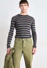 Stripete langermet skjorte i marineblått og beige med rund hals, sammen med olivengrønne bukser. En jakke holdes i venstre hånd.