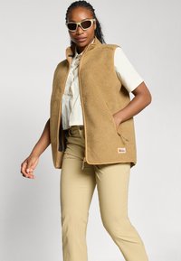 Femme portant une veste polaire beige sur une chemise blanche à manches courtes, pantalon beige et lunettes de soleil crème, debout devant un fond clair uni.