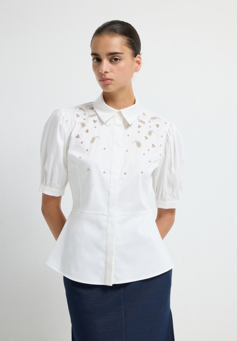 Witte blouse met pofmouwen, versierd met gouden versieringen. Structuurmatig passend, met een klassieke kraag en een knoopsluiting aan de voorkant.