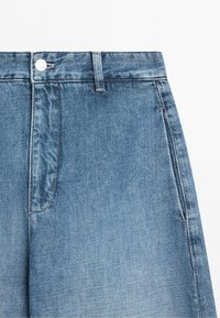 Niebieskie jeansy z denimu pokazujące pas, szlufkę na pasek, guzik, przedni suwak i przednią kieszeń na jednolitym białym tle.