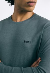 Koszulka z długim rękawem z okrągłym dekoltem w ciemnozielonym, prążkowanym materiale o teksturowej powierzchni. Posiada małe czarne logo "BOSS" na piersi.
