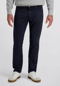 Mann in dunkelblauen Jeans, einem hellgrauen Sweatshirt und weißen Sneakers mit braunen Sohlen, der vor einem einfarbigen Hintergrund steht.