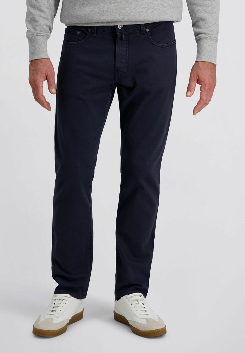 Mann in dunkelblauen Jeans, einem hellgrauen Sweatshirt und weißen Sneakers mit braunen Sohlen, der vor einem einfarbigen Hintergrund steht.