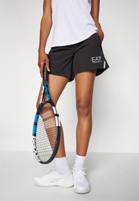 Sorte idrettsshorts med en logo, kombinert med en hvit T-skjorte og hvite tennissko. Holder en svart og blå tennisracket.