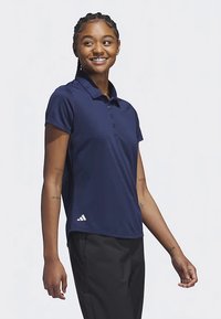 Νεαρή γυναίκα με πλεξούδες, που φοράει μπλε σκούρο πόλο μπλούζα και μαύρο παντελόνι, χαμογελά και κοιτάζει προς το πλάι σε απλό φόντο.