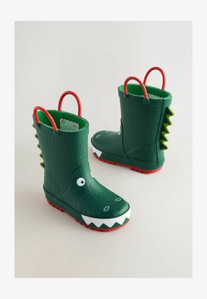 Zöld gumicsizmák krokodilbőr mintával, fehér fogakkal, nagy szemekkel, piros fogantyúkkal és piros talppal; tüskés díszek jellemzik a hátulját.