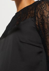 Gros plan sur un chemisier noir avec un empi ècement et des manches en dentelle florale transparente, corps en tissu lisse, porté par une personne à la peau brun clair.