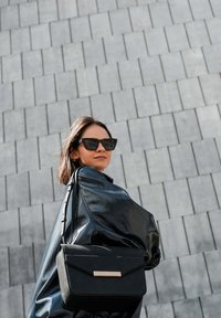 Zwarte leren handtas met een gladde, gestructureerde vormgeving en een gouden accent. Model draagt een glanzende zwarte jas en donkere zonnebril tegen een gestructureerde grijze achtergrond.