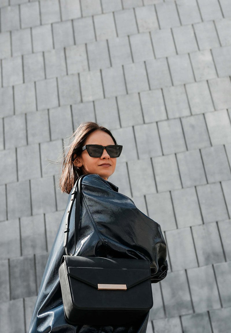 Zwarte leren handtas met een gladde, gestructureerde vormgeving en een gouden accent. Model draagt een glanzende zwarte jas en donkere zonnebril tegen een gestructureerde grijze achtergrond.
