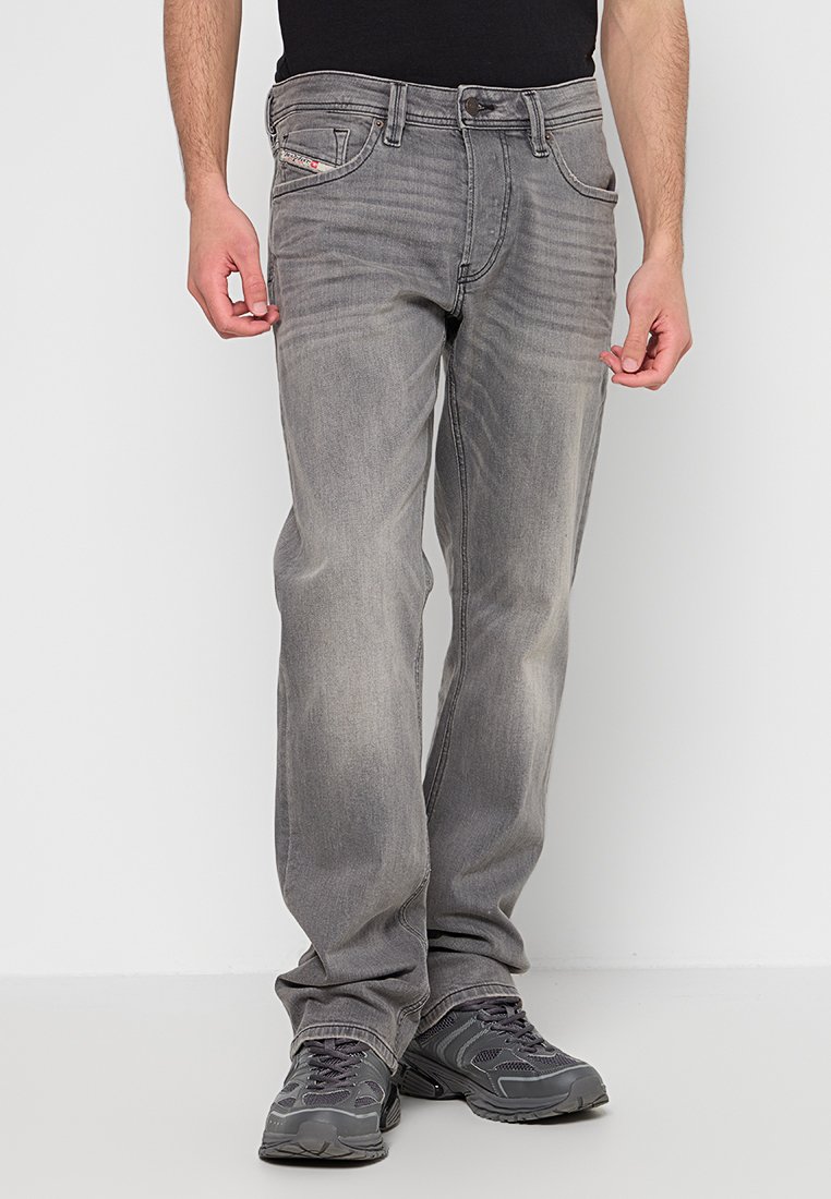 Mann in verblichenen grauen Jeans und dunkelgrauen Sneakers, der vor einem schlichten weißen Hintergrund steht, mit entspannten Händen an den Seiten.