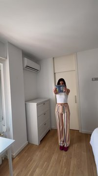 Νεαρή γυναίκα με ριγέ παντελόνι και λευκό τοπ στέκεται σε φωτεινό δωμάτιο κρατώντας ένα συσκευασμένο προϊόν προς την κάμερα, κοντά σε ένα λευκό κομοδίνο και κλιματιστικό.