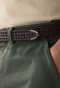 Ceinture en cuir tressé brun foncé avec boucle argentée, portée sur un tissu beige clair, associée à un pantalon vert texturé.