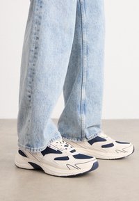 Zapatillas deportivas con una combinación de colores beige y marino, materiales de malla y ante, texturas combinadas y una suela blanca gruesa.