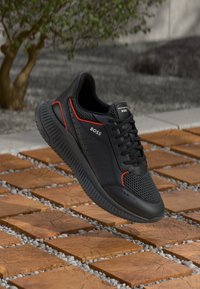 Chaussures de sport noires avec des panneaux en mesh, des accents rouges et une semelle texturée. Logo sur le côté, conception légère pour le confort.
