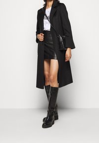 Zwarte lange jas met wijde mouwen, gecombineerd met een zwarte leren minirok en zwarte kniehoge laarzen. Kleine, gewatteerde zwarte tas als detail.
