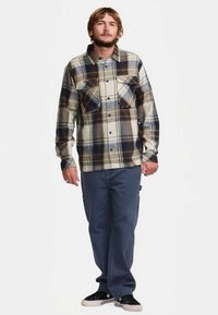 Camisa de flanela com um padrão de xadrez em tons de bege, azul e castanho escuro; dois bolsos no peito; gola; combinada com calças azuis de corte largo.