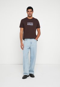 Tricou maro din bumbac cu text alb "Calvin Klein New York City", asortat cu blugi drepți albastru deschis și pantofi negri. Fundal simplu.