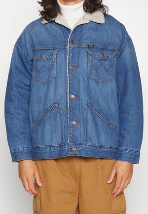 Persona que lleva una chaqueta azul de mezclilla con cuello de borrego sobre un suéter de cuello alto blanco, combinada con pantalones cargo marrones, de pie contra un fondo liso.