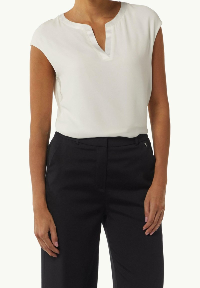 Femme portant un chemisier blanc sans manches avec un décolleté en encoche et un pantalon noir taille haute sur mesure, debout les mains détendues.