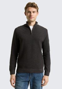 Dunkelgrauer Pullover mit halbem Reißverschluss, ausgestattet mit einem strukturierten Muster und gerippten Bündchen sowie Saum. Getragen über einem weißen Hemd, kombiniert mit blauen Jeans.