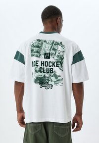 Camiseta de algodón blanca con detalles verdes, que presenta un gráfico retro del "Club de Hockey sobre Hielo" y un texto grande en la parte posterior. Manga corta, corte relajado.