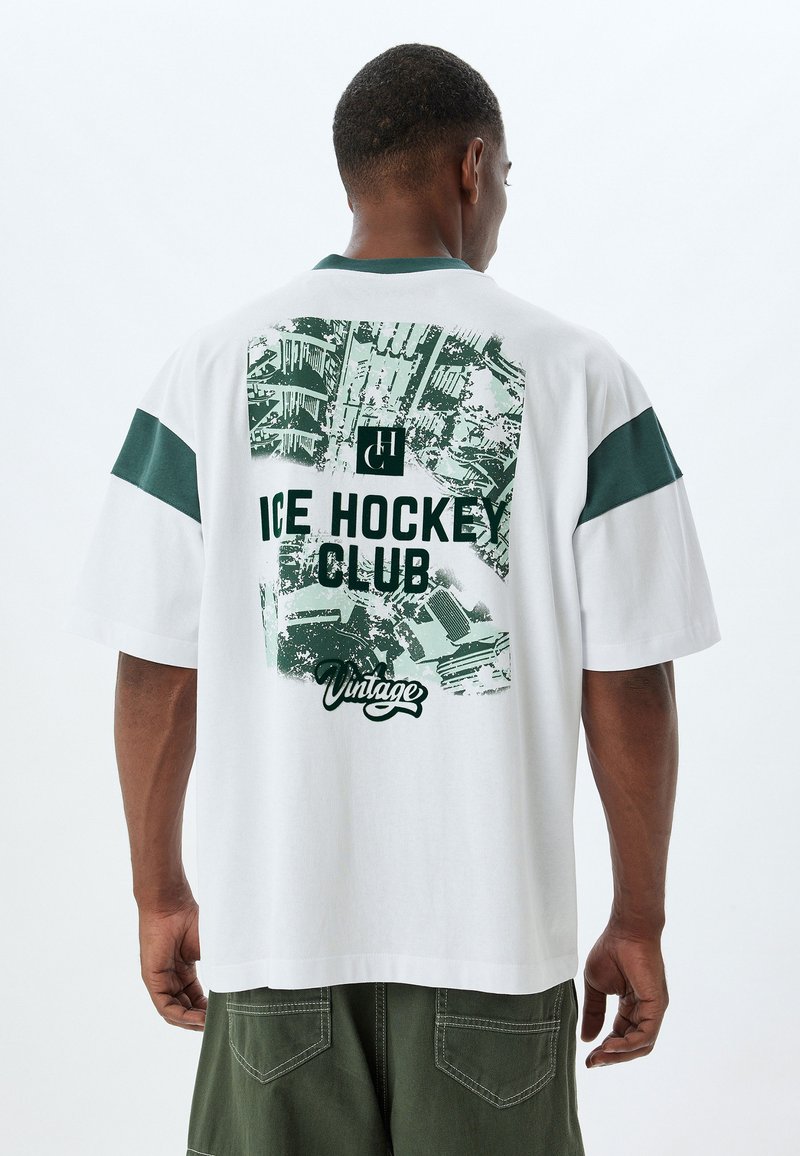 Camiseta de algodón blanca con detalles verdes, que presenta un gráfico retro del "Club de Hockey sobre Hielo" y un texto grande en la parte posterior. Manga corta, corte relajado.
