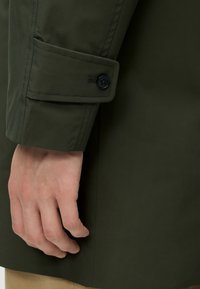 Αριστερό χέρι δίπλα σε μανίκι σκούρου πράσινου παλτό με λεπτομέρεια λουράκι με κουμπί, μπεζ παντελόνι μερικώς ορατό στο κάτω μέρος.