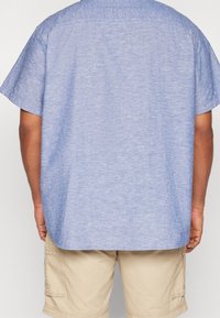 Světle modrá košile s krátkými rukávy a volným střihem, texturovaná látka a zakřivený lem. V kombinaci s béžovými cargo šortkami.