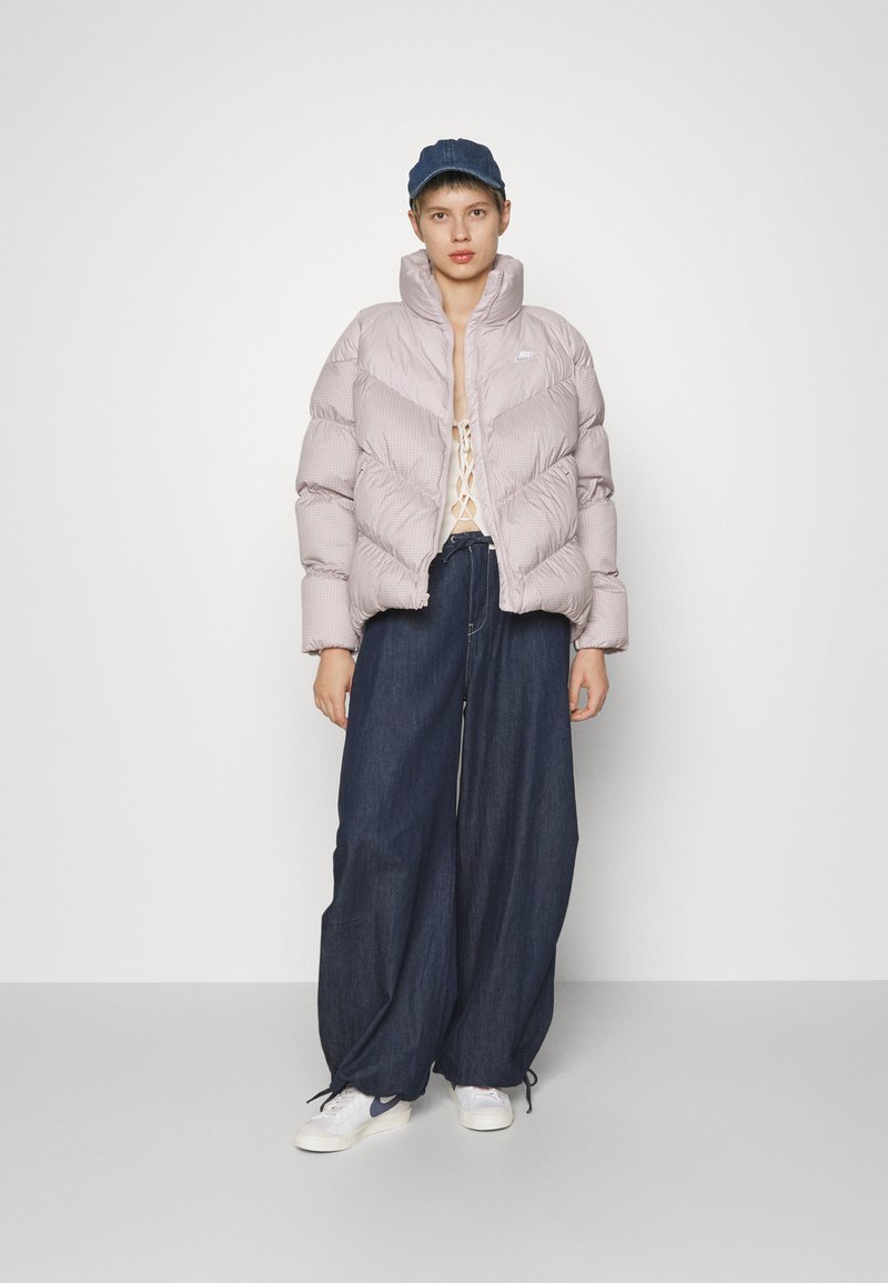 Doudoune rose clair avec un col haut, arborant un design matelassé ; associée à un pantalon en denim foncé ample et à des baskets blanches.