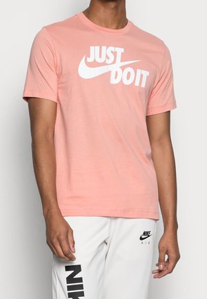 Jasnoróżowa bawełniana koszulka z okrągłym dekoltem, z dużym białym napisem "JUST DO IT" i logo Nike w kształcie łyżwy. Gładka faktura, krótkie rękawy.