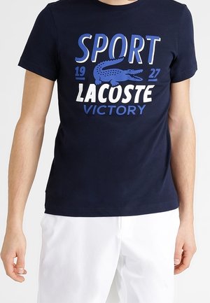 Granatowy t-shirt z napisem „SPORT 1927 LACOSTE VICTORY” i niebieskim logo krokodyla, noszony z białymi spodniami na osobie.