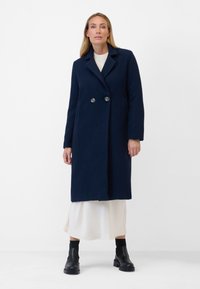 Mörkblå ullblandad kappa med texturerad finish, dubbelknäppta knappar och slag, bärs över en vit klänning. Svarta ankle boots.