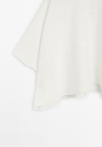 Vêtement blanc fluide avec un large ourlet et un design en couches, suspendu contre un fond clair, mettant en valeur sa texture douce et sa drape.