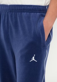 Tengerészkék melegítőnadrág sima felülettel, rugalmas derékrésszel, oldalsó zsebekkel és fehér hímzett Jumpman logóval a bal combon.