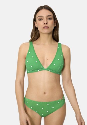 Mujer con bikini verde de triángulos con lunares blancos, de pie contra un fondo claro y liso, mirando al frente con expresión neutra.