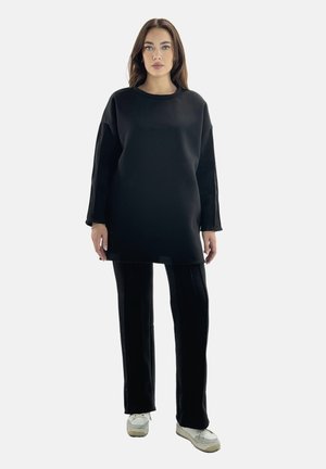 Ung kvinde står med front mod, iført en lang sort oversized sweatshirt, sorte bukser og hvide sneakers på en ensfarvet lys baggrund.