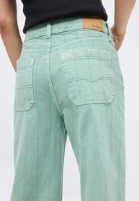 Οπίσθια όψη ατόμου που φορά ανοιχτό πράσινο τζιν με ραμμένες κάθετες ρίγες και δύο πίσω τσέπες, συνδυασμένο με μαύρο τοπ.