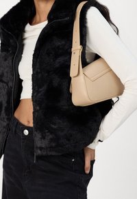 Beige strukturierte Handtasche mit rechteckiger Form und breitem Tragegurt, gehalten von einer Person, die eine schwarze Kunstfellweste und ein weißes Langarmshirt trägt.