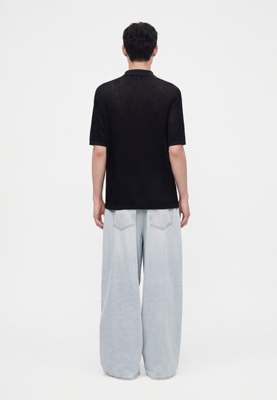 Homme portant une chemise noire à manches courtes et un jean bleu clair à jambes larges, debout, dos à un fond uni.