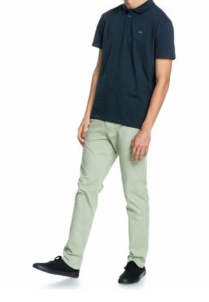Marineblauw poloshirt met kraag en groen logo, gecombineerd met lichtgroene slim-fit broek en zwarte sneakers. Casual outfit met nadruk op kleur en pasvorm.