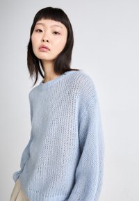 Pull en tricot bleu clair à coupe ample, avec un col côtelé et une texture en tricot ajouré, sur fond uni.