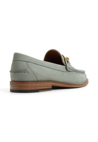 Mocasin de cuero verde claro con punta redondeada, pequeño tacón y suela de madera marrón en contraste; cuenta con un acento decorativo dorado.