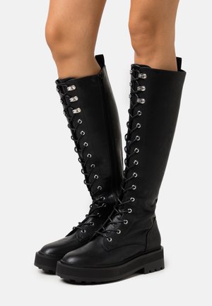 Bottes noires montantes à lacets avec semelles épaisses portées sur des jambes nues sur fond blanc.
